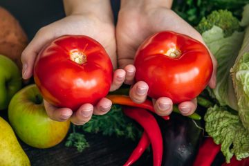Женские руки держат 2 спелых томата на фоне стола, на котором лежат другие овощи