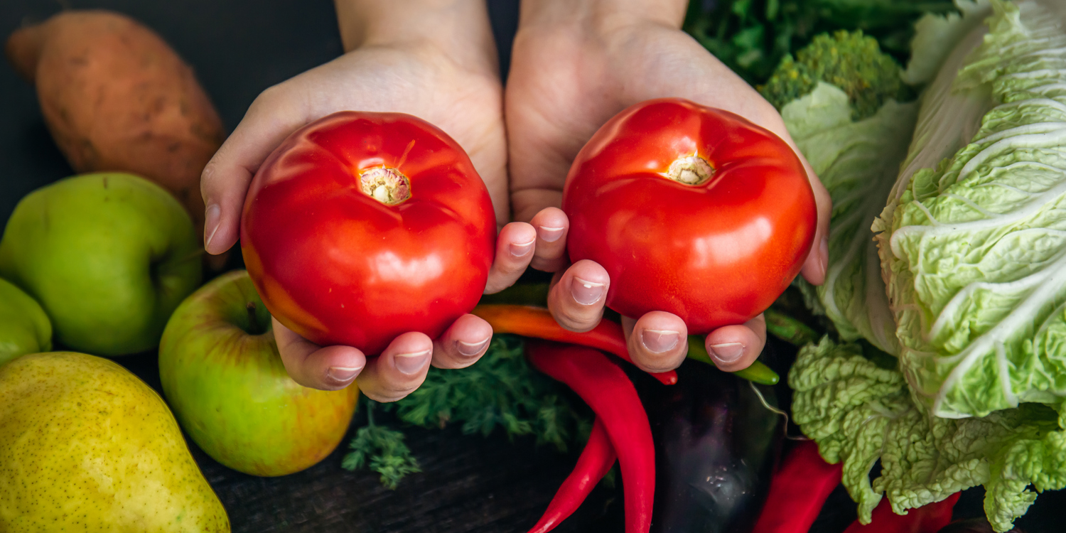 Женские руки держат 2 спелых томата на фоне стола, на котором лежат другие овощи