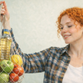 Рыжая девушка в белой футболке и клетчатой рубашке держит авоську с продуктами: кабачок, бутылка подсолнечного масла, зелёные яблоки, помидоры, лимон.