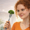 Рыжая кудрявая девушка держит в руке вилку с наколотым соцветием брокколи