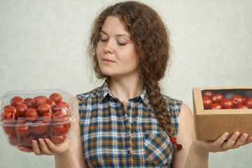 Девушка в клетчатой рубашке держит в одной руке пластиковый контейнер с помидорами черри, а в другой - картонную коробку с помидорами черри и сравнивает их
