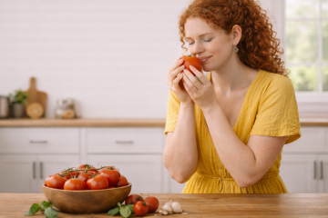 Девушка в желтом платье сидит на кухне с помидором в руках. Перед ней на столе миска с помидорами, рядом лежит еще несколько томатов, чеснок и базилик