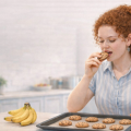 Кудрявая рыжеволосая девушка на кухне пробует печенье. На столе — противень с печеньем и связка бананов