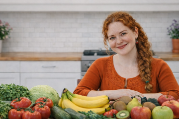 Женщина сидит на кухнеза столом. На столе лежат Бананы, огурцы, помидоры, киви, груши, яблоки