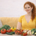 Молодая женщина в жёлтом платье сидитн адиване. перед ней на столе лежат овощи и фрукты: брокколи, помидоры, огурцы, жёлтый перец, шампиньоны, клубника
