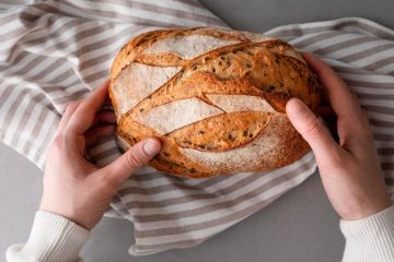 Человек в белом свитере тянется к свежей буханке хлеба, лежащей на полосатом полотенце