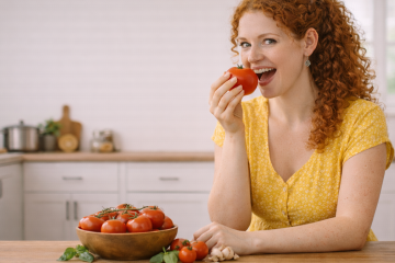Рыжая кудрявая девушка в желтом платье сидит на кухне за столом. На столет Стоит деревянная миска со спелыми помидорами, рядом лежит чеснок и базилик. Девушка держит в руке помидор и готовится откусить