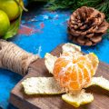 На столе ледат зелёные мандарины с листьями, ёлочные ветки, шишка и разделочная доска с почищенным мандарином