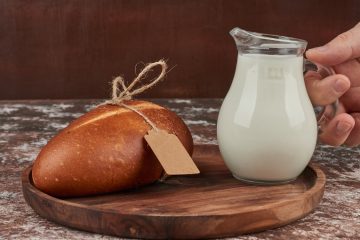 На столе стоит круглое блюдо, на котором лежит хлеб с картонной этикеткой. Человек ставит рядом стеклянный кувшин с кефиром