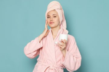 Молодая девушка в розовом халате и с замотанными в розовое полотенце волосами наносит на лицо крем