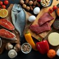 На темной поверхности лежат продукты: рыба, мясо, сыр, желтый и красный перец, яйцо, шампиньоны, зеленые яблоки, томаты черри на ветке, половина лимона, приправы и специи, сыр, молоко в пластмассовой бутылке, грейпфрут, грецкие орехи, хлеб