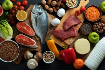 На темной поверхности лежат продукты: рыба, мясо, сыр, желтый и красный перец, яйцо, шампиньоны, зеленые яблоки, томаты черри на ветке, половина лимона, приправы и специи, сыр, молоко в пластмассовой бутылке, грейпфрут, грецкие орехи, хлеб