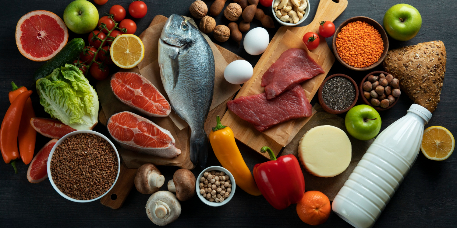 На темной поверхности лежат продукты: рыба, мясо, сыр, желтый и красный перец, яйцо, шампиньоны, зеленые яблоки, томаты черри на ветке, половина лимона, приправы и специи, сыр, молоко в пластмассовой бутылке, грейпфрут, грецкие орехи, хлеб