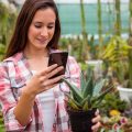 Девушка в клетчатой рубашке фотографирует небольшое растение в горшке на телефон