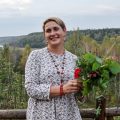Женщина в светлом платье стоит у деревянного забора с букетом трав