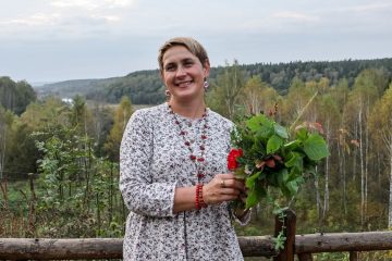 Женщина в светлом платье стоит у деревянного забора с букетом трав