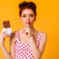 Молодая женщина в белом платье в красный горошек держит в руке плитку шоколада, а указательный палец другой руки прижимает к губам