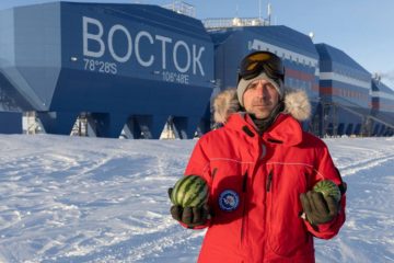 Мужчина в красном пуховике стоит на фоне полярной станции "Восток" с двумя арбузами в руках