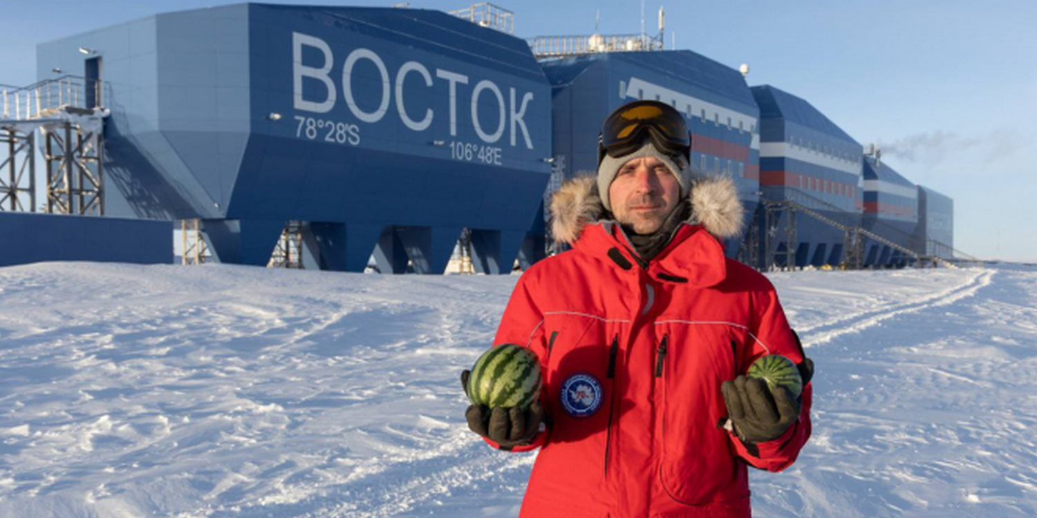 Мужчина в красном пуховике стоит на фоне полярной станции "Восток" с двумя арбузами в руках