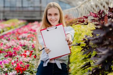 Девушка стоит в промышленной теплице, в которой выращивают цветы и держит в руках планшет с белым листом бумаги