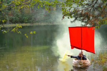Женщина плывет по озеру в лодке, выдолбленной из гигантской тыквы с красным парусом и держит в руке дымовую шашку