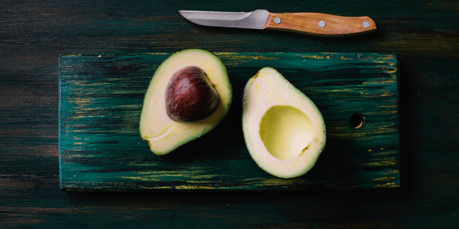 На разделочной доске лежит разрезанный на половинки авокадо. Рядом лежит нож