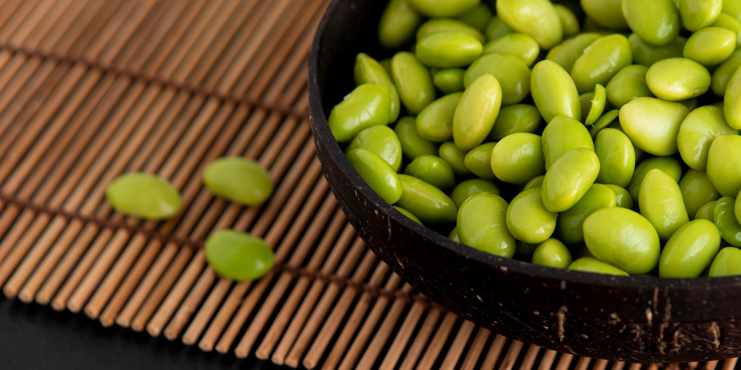 На бамбуковой салфетке стоит круглая пиала со свежими бобами эдамамэ. Три боба выпали и лежат рядом