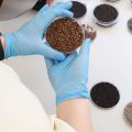 На столе лежит несколько чашек Петри с различными семенами. У стола стоит человек в белом халате и одержит одну из чашек Петри в руках