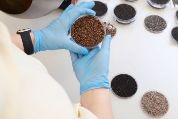 На столе лежит несколько чашек Петри с различными семенами. У стола стоит человек в белом халате и одержит одну из чашек Петри в руках