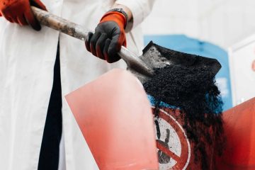 Рабочий в белом халате и защитных руковицах совковой лопатой пересыпает биоуголь